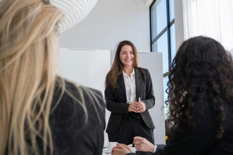 A woman wearing a suit stands at the front of a room while speaking to other people