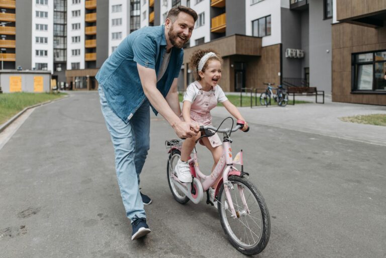 A man holds a bicycle while his young daughter rides it