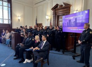Several people sit in front of a large group of police and one stands at a podium.